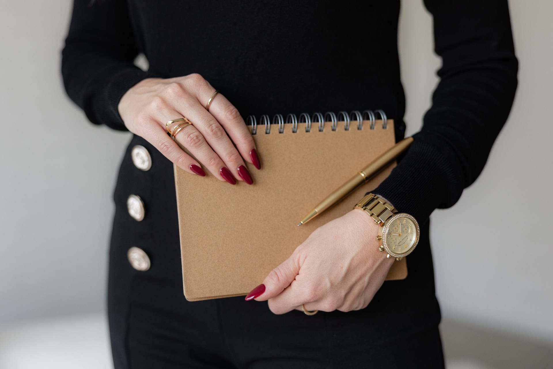 Gepfelgte Frauen Hand mit rot lackierten Nägeln, die einen Terminkalender in der Hand hält. Sie trägt ein schwarzes Outfit und goldenen Schmuck.